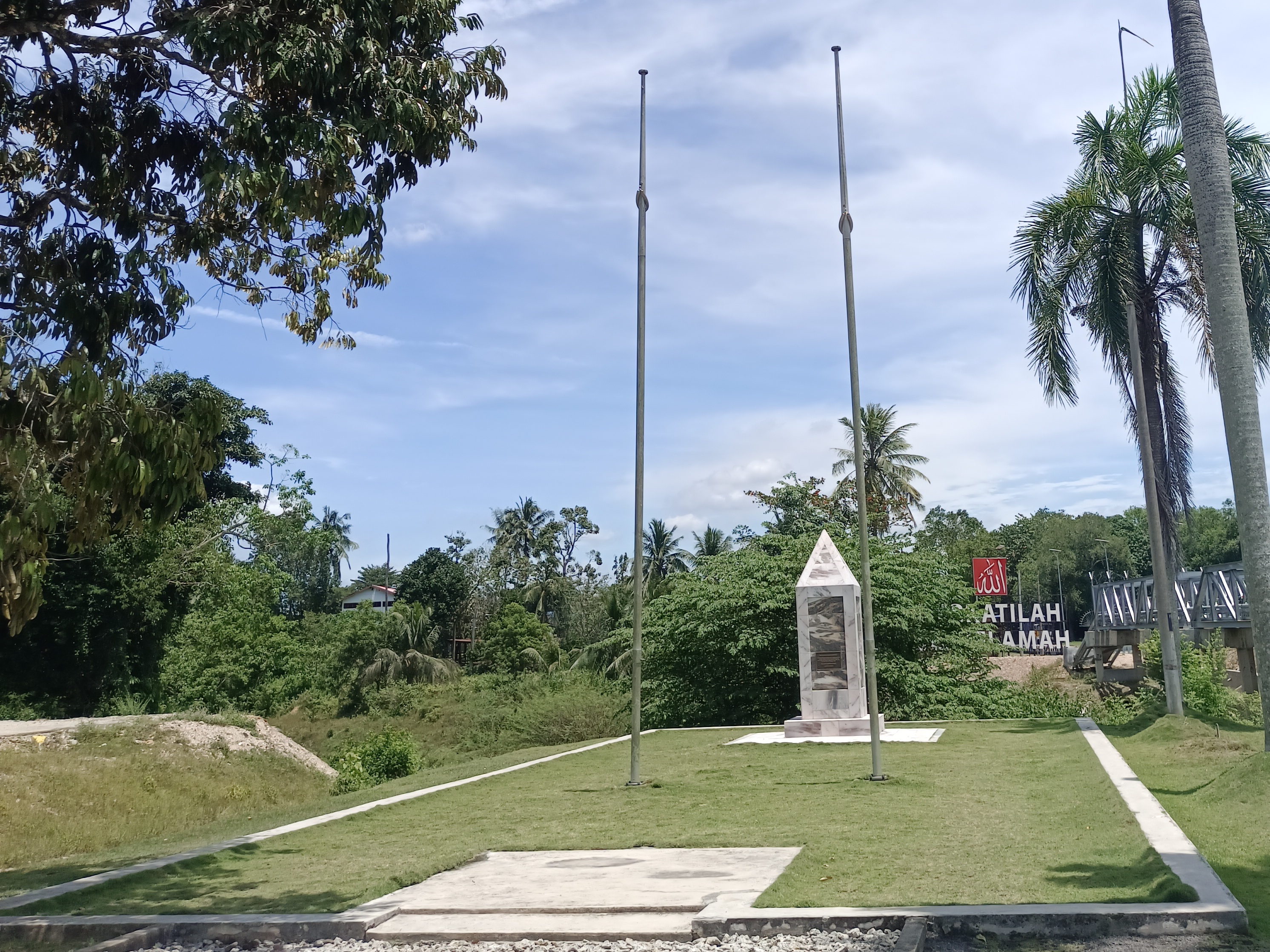 The flagpoles and monument of the Kelamah River Memorial facing westwards (with the Allah Berkatilah Sungai Kelamah sign at the background)
