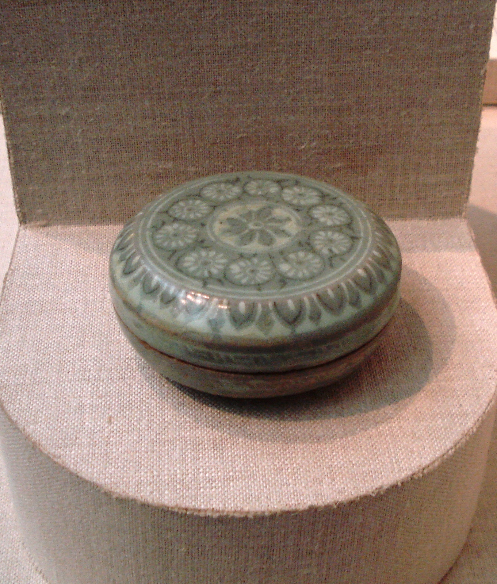 A Korean stoneware cosmetic box with white and black inlay and a celadon glaze, from the Goryeo Dynasty, dated c. 1150–1250. Items such as these have been excavated from Korean Goryeo tombs alongside ceramic hair-oil bottles, needle cases, bronze mirrors, and hairpins.