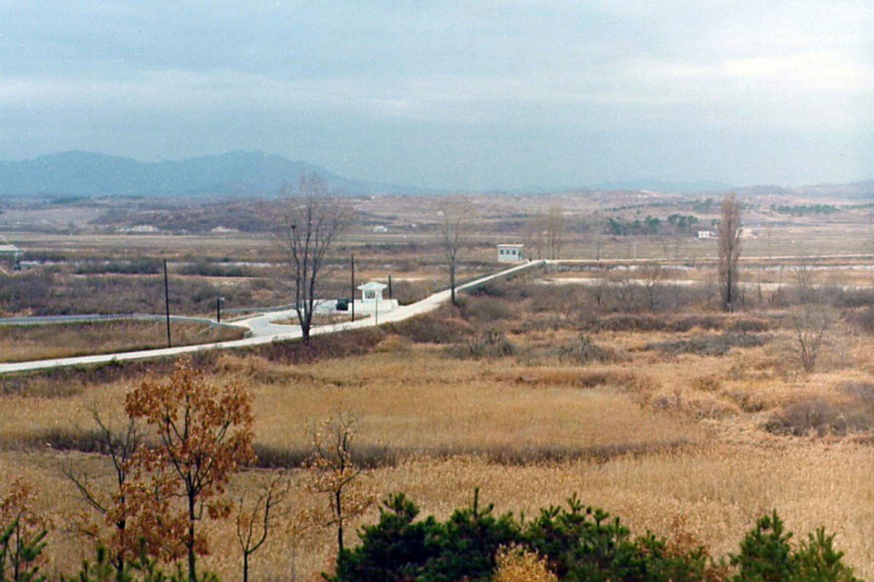 I took this picture in December of 1975, from the same location the picture of the Axe Murder Incident were taken from 8 months later. At the closest end of the Bridge of No Return is CP#3, "The Lonliest Outpost in the World". At the furthest end of the bridge is North Korean territory, outside of the JSA. Notice the 2 1/2 ton truck sitting at CP#3. The two people who would work there during daylight hours needed a big, heavy truck to break through the road barriers the North Koreans had erected for the occasions when they tried to block the road. Also visible in the picture is the tree the fight was over. In summertime, the tree was twice as full as it appears in this picture, basically obscuring visibility between UNC posts. Since the North Koreans would try to kidnap the people working at CP#3 and drag them across the bridge, maintaining visibility between the checkpoints was imperative for their safety during the summer months.