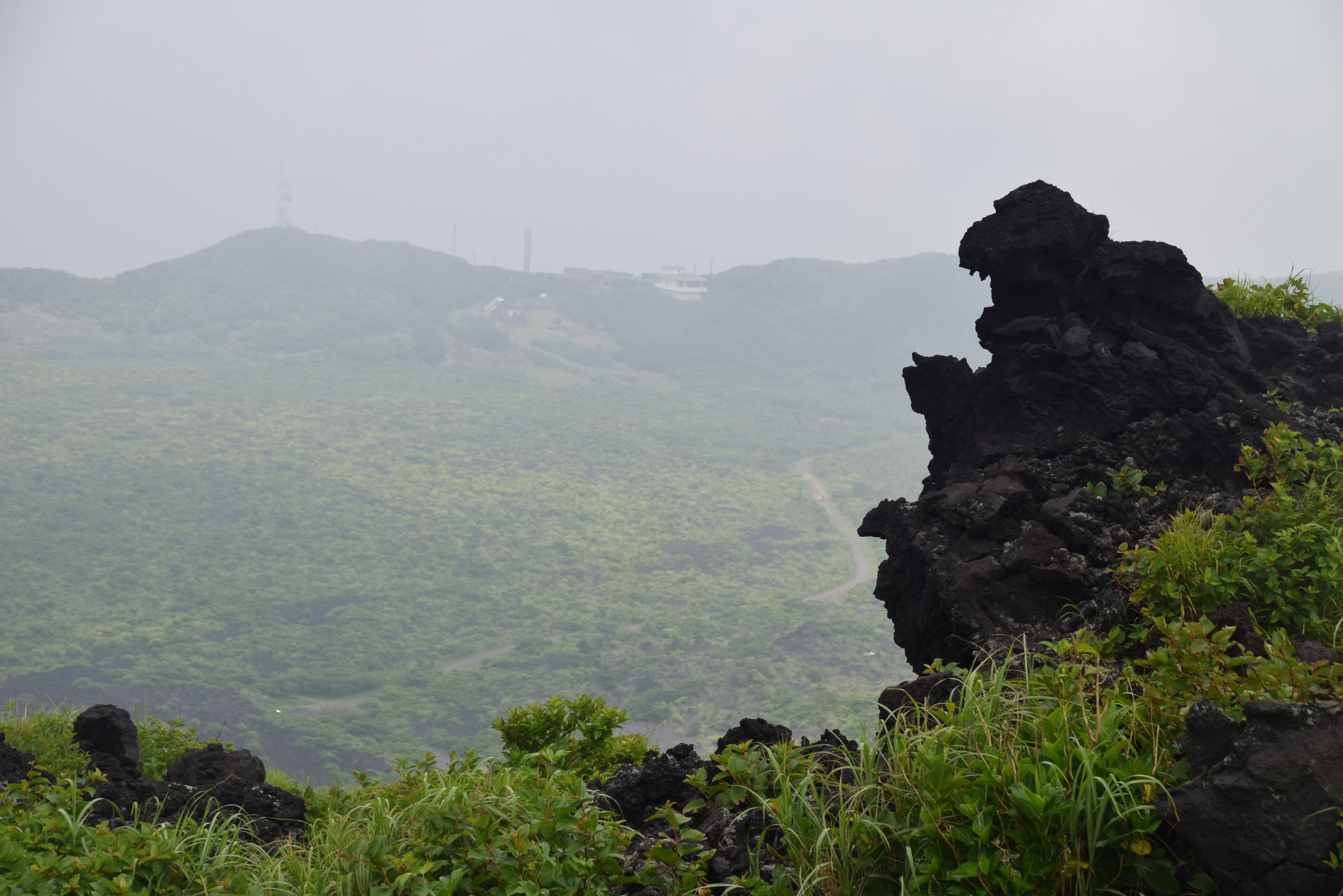 Godzilla Rock (Ja: ゴジラ岩) is located on the inner caldera rim path (火口一周道路) around the crater of Mount Mihara (三原山), on Izu Oshima (伊豆大島), Japan. In the back can be seen the paved walking path from the bus stop on the outer caldera rim to the top of the mountain.