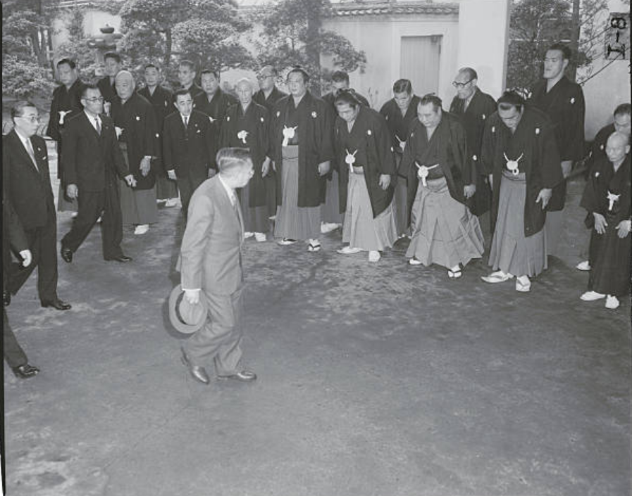 Emperor Hirohito being greeted by officials and Sumo grand champions at the entrance of the Kuramae Kokugikan Hall.