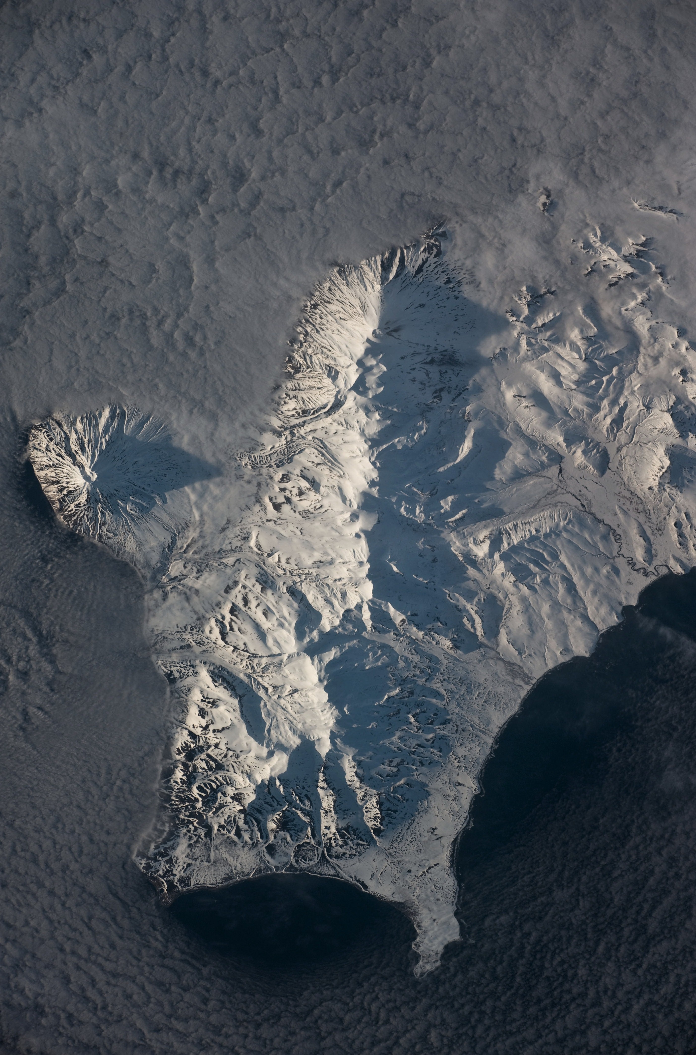 This astronaut photograph shows the southern end of Paramushir Island after a snowfall. The western slopes of the mountains are brightly illuminated, while the eastern slopes are in shadow.
Four major volcanic centres create this part of the island. Fuss Peak (image centre left) is an isolated stratovolcano connected to the main island via an isthmus. Fuss Peak last erupted in 1854.
The southern tip of the island is occupied by the Karpinsky Group of three volcanic centres. A minor eruption of ash following an earthquake occurred on this part of the island in 1952.
The Lomonosov Group to the north-east (image centre) includes four cinder cones and a lava dome that produced several lava flows in the past, but there have been no eruptions from the Lomonosov Group in recorded history.
The most recent volcanic activity on Paramushir Island occurred in 2008 at the Chikurachki cone located along the northern coastline of the island at image top centre. The summit of this volcano [1,816 meters above sea level] is the highest on Paramushir Island.

Much of the Sea of Okhotsk visible in the image is covered with low clouds that often form around the islands in the Kuril chain. The clouds are generated by moisture-laden air passing over the cool sea/ocean water, and they typically wrap around the volcanic islands.