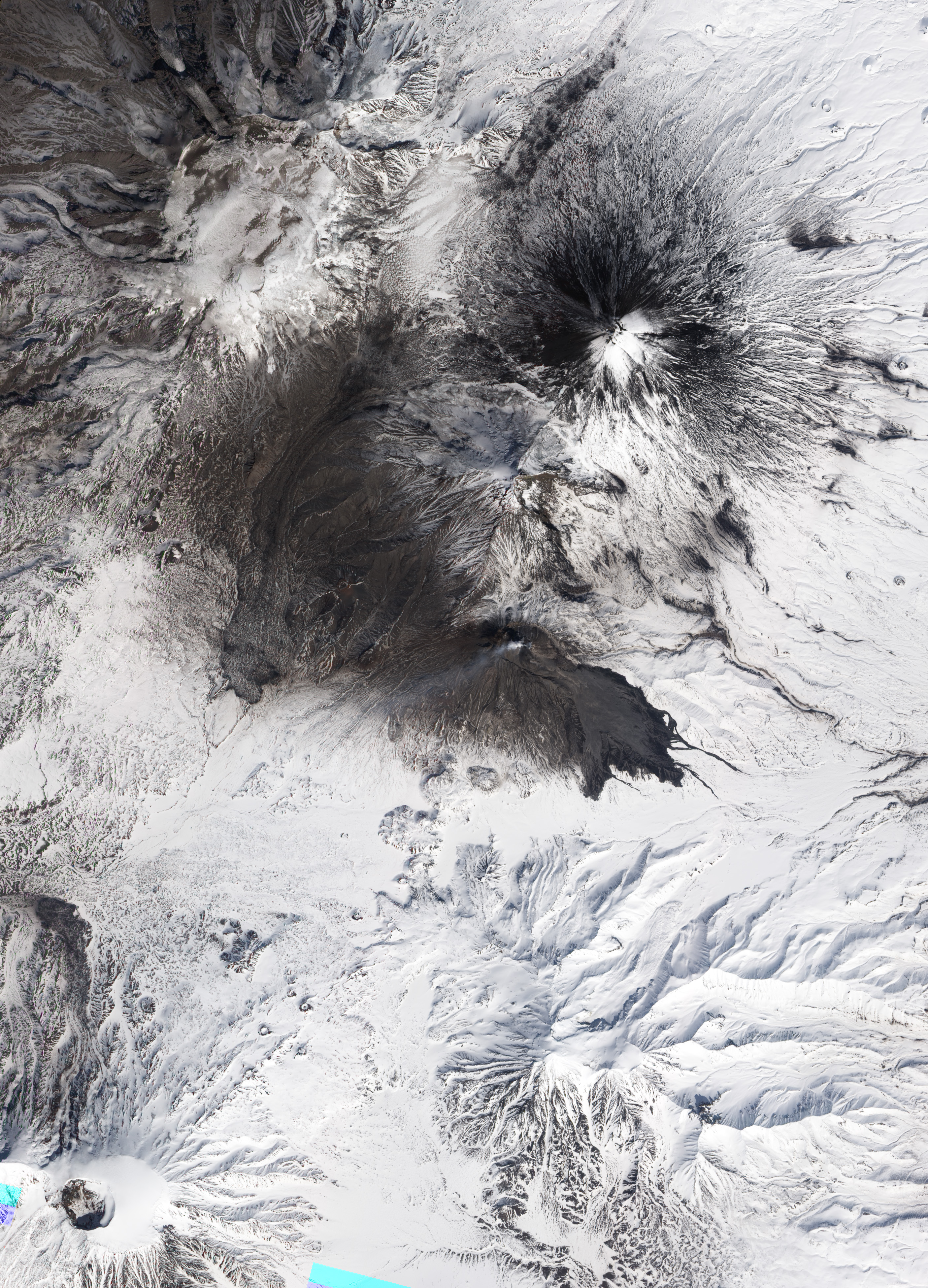 Natural-colour satellite image showing evidence of an eruption at Bezymianny Volcano. Dark volcanic deposits (likely a combination of pyroclastic flows and lahars) extend more than 7.3 kilometres into valleys to the south-east. A light-coloured plume of ash, steam, and sulphur dioxide rises above Bezimianny’s summit and blows towards the west. Volcanic ash covers the upper slopes of the volcano, especially to the south and west. White snow, still deep in late April, blankets the surrounding landscape.
