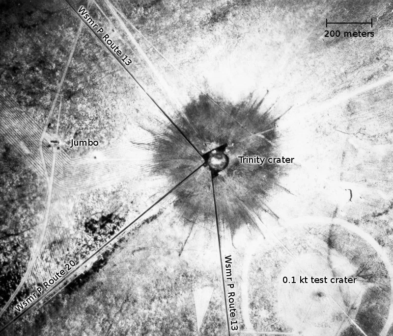 White Sands, New Mexico. Aerial view of the aftermath of the Trinity test, 28 hours after the explosion. The smaller crater to the southeast is from the earlier detonation of 100 tons of TNT on May 7, 1945. To the left of the crater can be seen the "Jumbo" container, unharmed, and its collapsed tower (a vertical line).