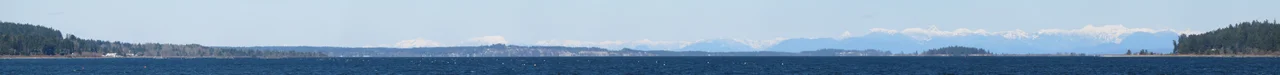 Panoramic combination of 14 images taken from parking area behind Petro Canada service station above BC Ferries Terminal at Buckley Bay, British Columbia across Baynes Sound. Bluffs in the centre of the image are Cape Lazo. Near forest to the right is Denman Island across Baynes Sound. The snow covered peaks in the distance surround Desolation Sound across the Strait of Georgia.