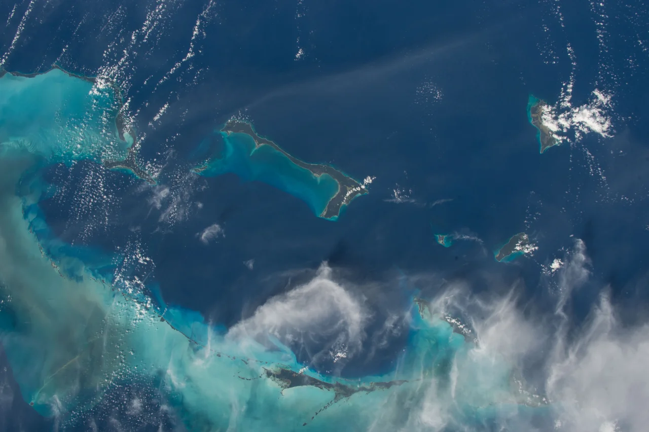 Like most other islands in the Bahamas, Cat Island is located on a large depositional platform that is composed mainly of carbonate sediments and surrounding reefs. The approximately 77 kilometre-long island is the part of the platform continuously exposed above water, which allows for soil development (brown to tan areas) and the growth of vegetation.
Shallow water to the west-south-west (below the island in this view) appears bright blue, in contrast to the deeper ocean waters to the north, east, and south. In this astronaut photograph, the ocean surface near the south-eastern half of the island has a slight grey tinge due to sun-glint, or light reflecting off the water surface back towards the International Space Station. Small white cumulus clouds obscure some parts of the island.