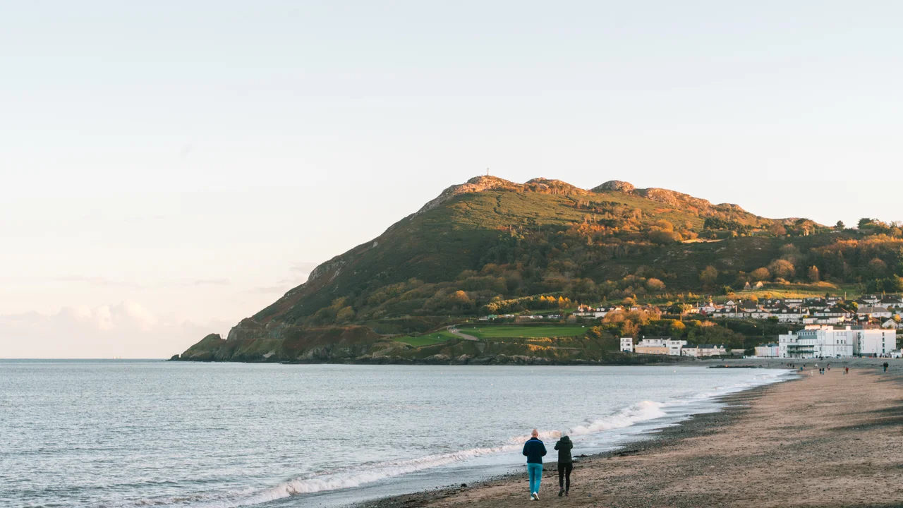 A view of Bray Head, County Wicklow, Ireland, seen from Bray's beach during autumn. The low evening sun illuminates the hillside. Two people are walking along the shingle beach in the foreground, with the Irish Sea to the left and seafront buildings visible at the base of the hill.