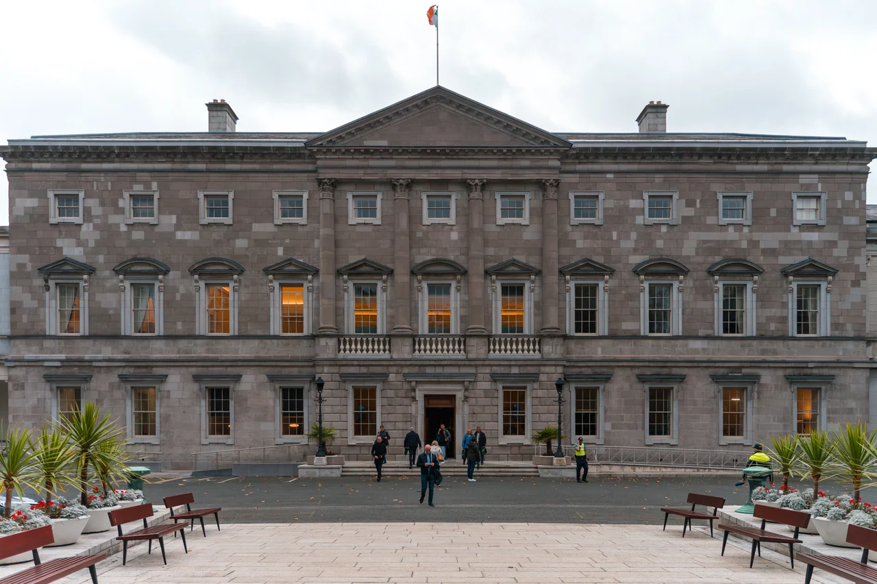 A head-on, symmetrical view of the main façade of Leinster House in Dublin, Ireland. The large, grey-stone neoclassical building, home to the Oireachtas (Irish parliament), is seen from its forecourt. Several people are visible walking near the main entrance at the centre. The Irish tricolour flag is flying from a flagpole on the roof. The foreground includes benches, potted plants, and paving stones.