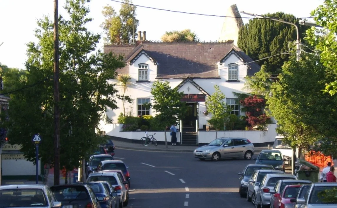 Part of Main Street, Raheny, Dublin, looking uphill towards a former school house (now a restaurant) which in turn backs on to the site of the old village church, (Old) St Assam's. The River Santry runs under the road in the foreground.