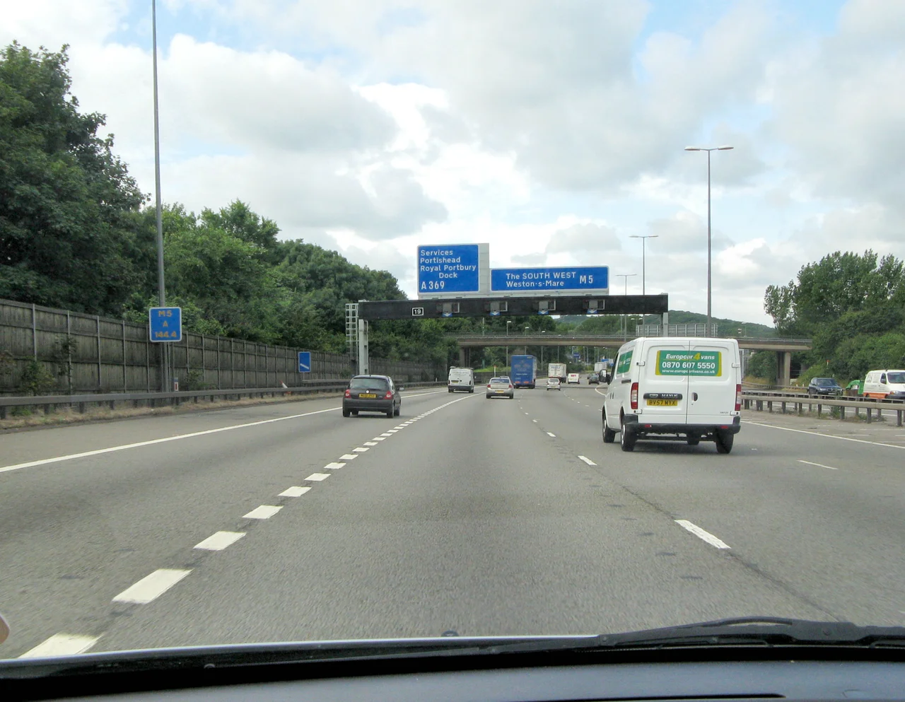 View from my car on the southbound M5 motorway approaching the Gordano Services, just south of the Avonmouth Bridge, Bristol, England. 
THE SLIP LANE on the left serves the Gordano Services and the A369 road, marked by the closely-dashed white dashes (with the dark car and white van on it) . The single diagonal line on a blue background indicates 100 metres to go before the exit (useful in fog or rain). (The carriageway markings are the well-spaced-out white dashes).
THE OVERHEAD GANTRY says “The SOUTH WEST” meaning the south west of England. The junction number is shown as 19 (the next exit would be 20 for Clevedon). Below the blue down-pointing arrows on the gantry crosses can be lit up to bar a carriageway.
THE BLUE NOTICE on the far left is not for the motorist, it helps maintenance, police, planners etc locate a point on the motorway. “A” means the southbound carriageway and “144.4” is a distance from a reference point. 

Photographed by Adrian Pingstone in July 2009 and placed in the public domain.