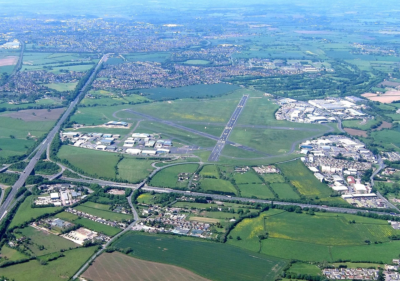 An aerial view of Gloucestershire Airport, Gloucestershire, England.Looking WEST from the open window of a landing Piper Cherokee Warrior (reg G-BBXW) during a pleasure flight from the airport.Running up and down the picture (on the left) is the straight A40 London to Fishguard road, and running across the picture (at the bottom) is the M5 motorway from Exeter to Birmingham.In the distance is Innsworth and then Gloucester.The aircraft was a Piper Cherokee Warrior (G-BBXW).