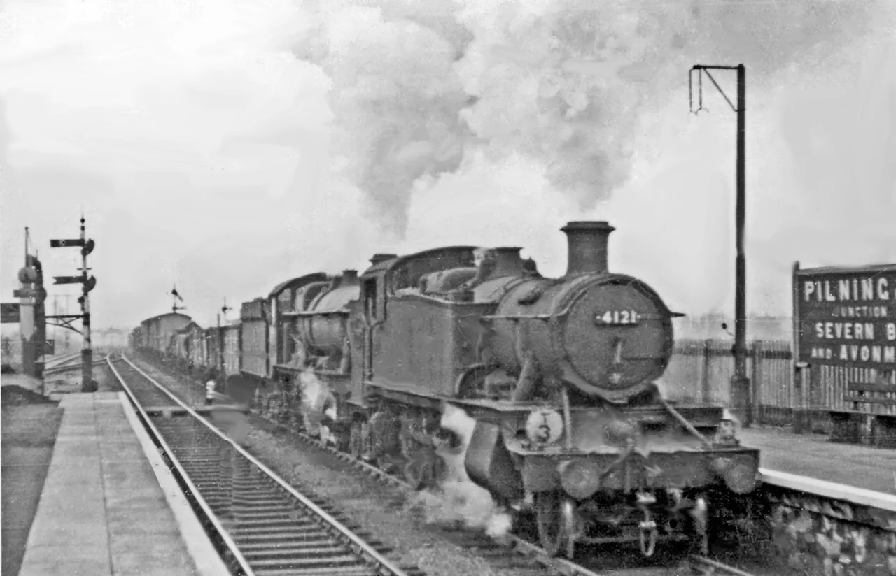 Piloted Up freight climbing out of the Severn Tunnel through Pilning High Level station.
One of the 2-6-2Ts dedicated to assisting freight trains through the Tunnel, Collett '5100' No. 4121 (built 12/37, withdrawn 6/65, but was preserved and is currently undergoing restoration at Tyseley Locomotive Works), is piloting Collett 4-6-0 No. 4998 'Eyton Hall' (built 3/31, withdrawn 10/63) - a fine sight and sound. Note the sign 'Pilning Junction for Severn Beach and Avonmouth': the service via Severn Beach was still in operation (until 11/64).
