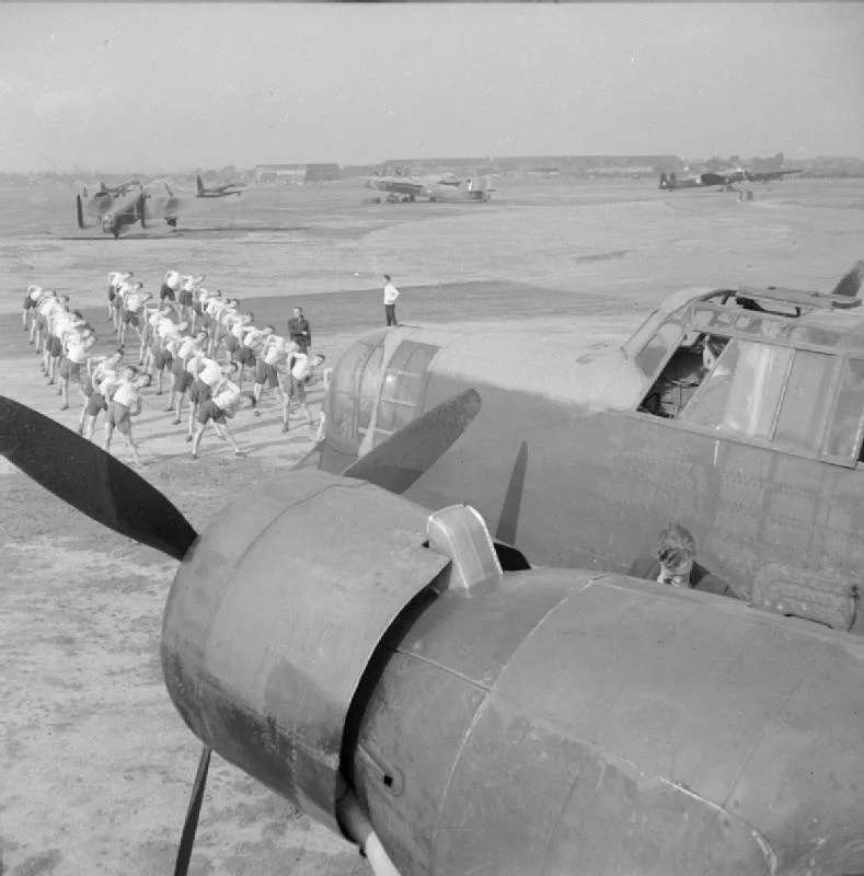 Paratroopers from the 6th Royal Welch Parachute Battalion undergoing physical training at Ringway, UK. In the foreground a Whitley III is serviced.