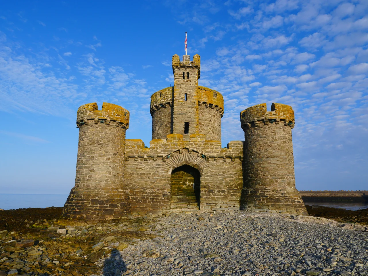 The Tower of Refuge stands upon St. Mary's Isle (also known as Connister Rock) - a marine hazard upon which many ships approaching Douglas Harbour wrecked due to low visibility or bad weather. The miniature castle was built in response to the 1830 destruction of the packet St. George with local donations. The project, undertaken by Sir William Hillary and designed by architect John Welch, was completed in 1832 and increased the visibility of the waterway hazard as well as provided a storage place for dry provisions in case anyone shipwrecked upon the isle. The islet is seen here at an average tide. Low tide allows one to walk out from the Douglas shore. Extremely high tide will completely submerge the islet from sight except for the Tower of Refuge.