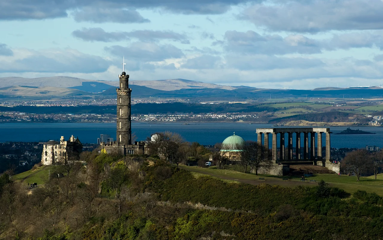 A view of Calton Hill from Salisbury Crag. The hill includes: the National Monument (Parthenon replica), the Nelson Monument (tall tower), the Old Observatory House (house on left) and the City Observatory (green dome). The Firth of Forth is the river (or Fjord if you prefer) in the background.