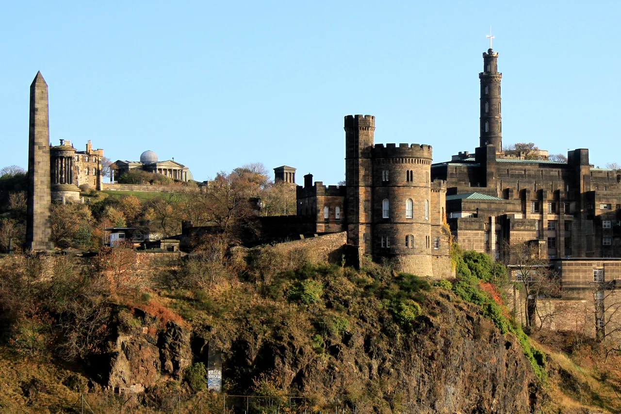 A view of Calton Hill from Salisbury Crag. The hill includes: the National Monument (Parthenon replica), the Nelson Monument (tall tower), the Old Observatory House (house on left) and the City Observatory (green dome). The Firth of Forth is the river (or Fjord if you prefer) in the background.