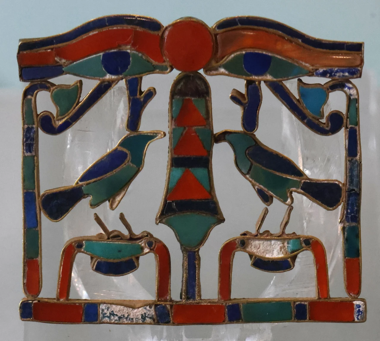 The English archaeologist Reginald Engelbach discovered this Ancient Egyptian pectoral jewellery in a tomb at Riqqeh, Egypt, in 1912 near the entrance to the Faiyum lake region. Its tomb ceiling had collapsed, crushing a tomb robber, which is probably why this jewellery survived.  This pectoral is a piece of golden chest jewellery with stones of carnelian, turquoise and lapis lazuli. This small (4.2cm wide) object has perhaps the most dramatic biography of any in the Manchester Museum collection, and one which would not seem out of place in a Hollywood movie script. Known today as the Riqqeh Pectoral after the site at which it was discovered, this ornate chest ornament, with two loops for suspension indicating that it was worn on a necklace, is an undoubted highlight of the Manchester Museum. The piece was created using a technique termed cloisonné, in which separate gold sections are filled with semi-precious stones. Lapis lazuli (dark blue), carnelian (red) and turquoise (blue/green) give the pectoral its colourful appearance and gem-like lustre. The reverse is chased in gold with details of the figures: two wedjat eyes (or ‘eyes of Horus’) flank a sun disk above two falcons (sometimes described as ‘crows’) on symbols for ‘gold’. The composition is arranged symmetrically around a stylised papyrus umbel suggesting a sekhem sceptre – a symbol of power. Two inward turned papyrus stalks frame the group.
Riqqeh pectoral. Manchester Museum Accession No 5966. 

The pectoral was found in association with two other items, each in the form of a king's name: Senusret II (Khakheperre) and Senusret III (Khakaure). It can, therefore, be reliably dated to the second half of the Twelfth Dynasty of Egypt (c.1900-1800 BC) of the Middle Kingdom of Egypt. It is a fine example of delicate jewellery on a small scale, typical of the best Middle Kingdom royal pieces.[1]