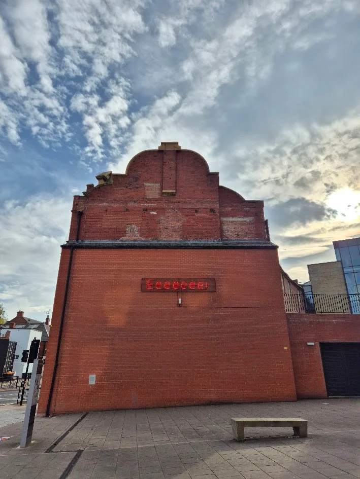 Mackem Shibboleth is a neon artwork displayed on the gable end of a red brick building. It is red neon characters showing the letters Eeeeeee