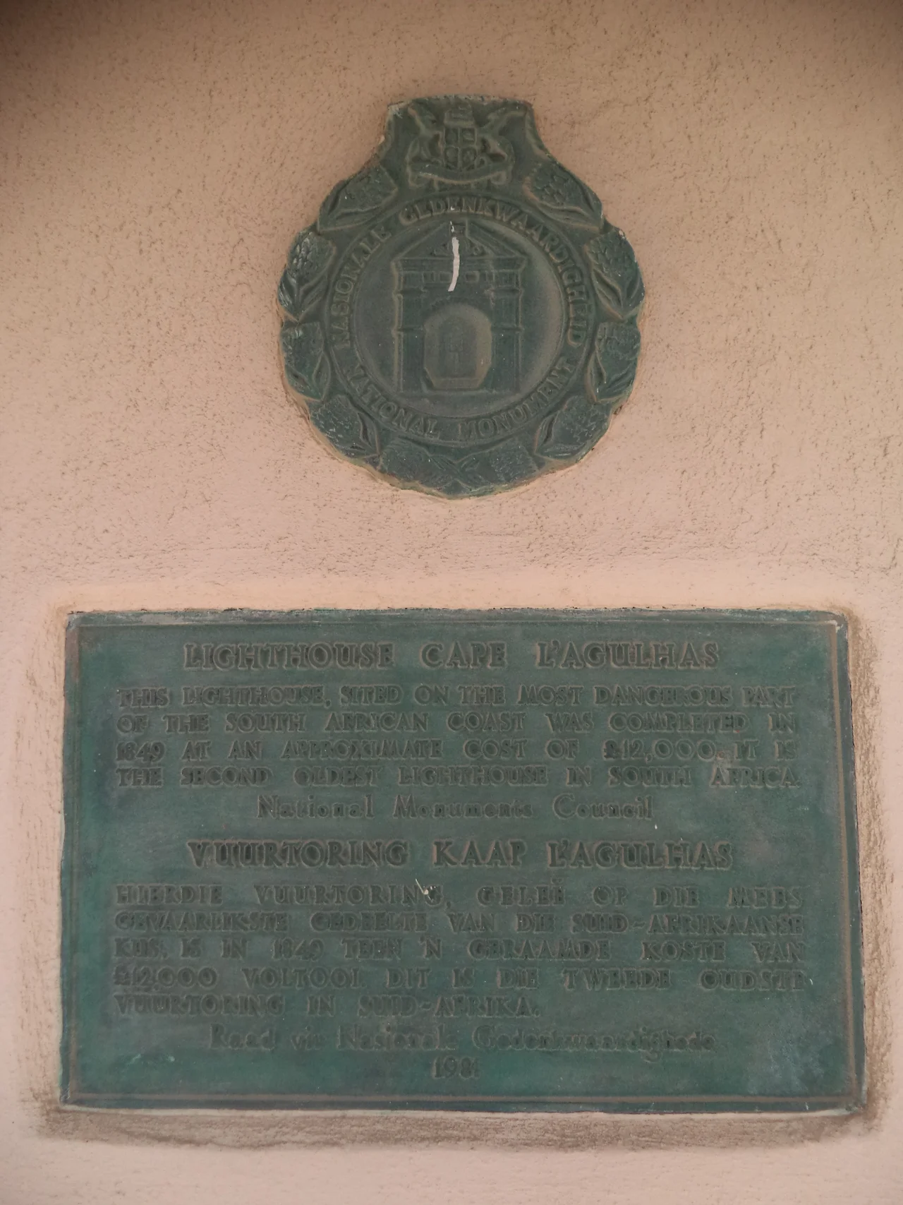 The memorial seal and the memorial plaque on the entrance of Cape Agulhas Lighthouse. The inscription on the seal writes:
National Monument
The inscription on the plaque writes:

Cape Agulhas Lighthouse 
This lighthouse, sited on the most dangerous part 
of the South African coast was completed in 
1849 at an approximate cost of £12,000. It is 
the second oldest lighthouse in South Africa. 
National Monument Council