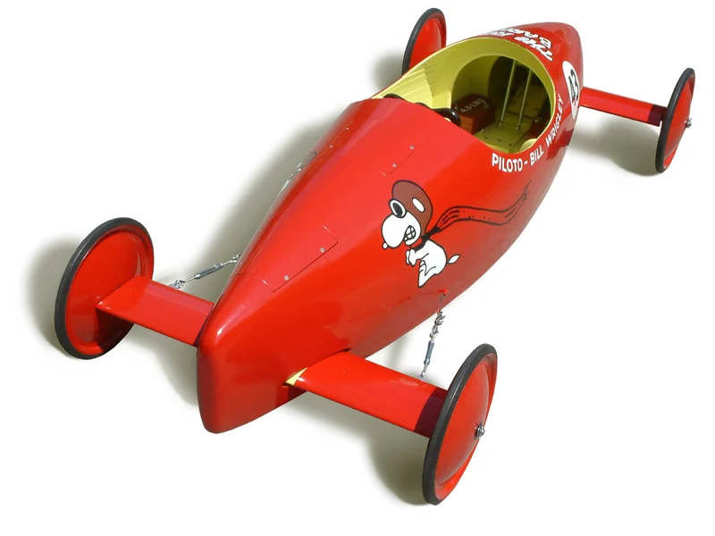 Image of a soap box derby racer of a late 1960s design, complete with the official Soap Box Derby issue wheels, painted red.
Seen here is an exact replica of a hand-built racer constructed in 1967 by Bill Wrigley Jr., supervised by his father, and raced the following year at the Kinsmen Coaster Classic, a soap box race in Montreal, Canada. It was raced for five years in total by Bill Jr. and younger brother Derek at various organized races throughout Quebec and Ontario.