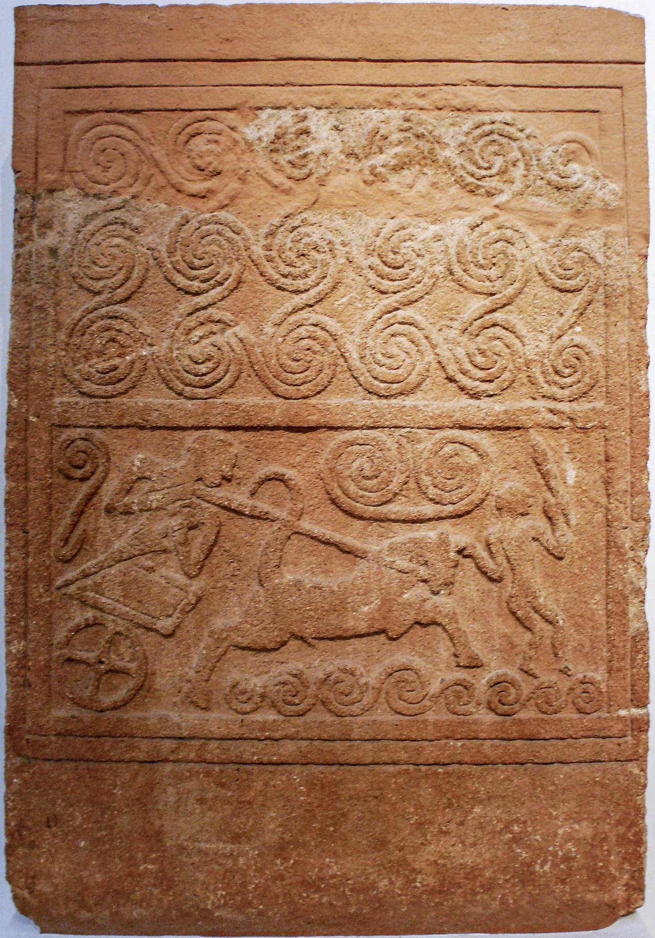 Funerary stele made of Poros stone with chariot scene relief. Mycenae, Grave Circle A, Grave V. 1500 BC.
National Archaeological Museum of Athens. The spirals in the upper panel may represent waves, thus indicating the coastal location of the scene in the lower panel. The standing charioteer pulls at the reins, while a second figure, who holds a weapon, possibly a sword, in his left hand, is depicted in front of the chariot.