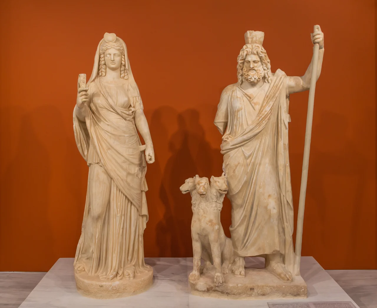 Group of ancient Roman statues of Persephone (as Isis), Cerberus, and Pluto (as Serapis), from Gortys. Archaeological Museum of Heraklion, Crete, Greece.These gods were worshiped in the Greek world from the Hellenistic period onwards. Pluto/Serapis has the modes on his head, a utensil used for the measurement of grain. Persephone/Isis with covered head, bears her symbols at the forehead (the crescent moon, the solar disk and the snake (uraeus). She is depicted holding in her right hand the sistrum, an egyptian musical instrument also known to Crete from prehistory, and in the left probably the straps that kept the dog Cerberus. The inclusion of Cerberus, guard of the underworld, in the group defines the two deities, despite their Egyptian symbols, as Pluto and Persephone, gods of the underworld. The group is a typical example of syncretism during Hellenistic and Roman times.