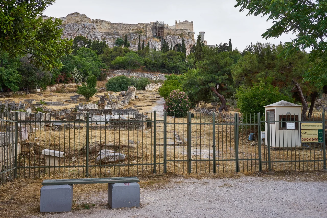 "In the southeast of the Ancient Agora in an area with impressive rock foundations, looms the "Eleusinion in the City" ("εν άστει Ελευσίνιο") between the Panathenaic Way on the west and the two branches of the "Street of the Tripods" on the north and south. It was a shrine sacred to the mystery religion of the goddess Demeter and Persephone along with their mortal counterpart Triptolemos. These Eleusinian deities were worshipped here already in the 6th century B.C., in an open-air shrine surrounded by a wall. In the second quarter of the 5th century B.C. the shrine received a small rectangular temple oriented north-south. Of this Ionic tetrastyle amphiprostyle temple, with an inner shrine and shallow pronaos are preserved only the groundwork of three sides. In the 4th century B.C. the need to reinforce the embankment of the temple led to the construction of a strong retaining wall. The area north of the temple embarkment, which is at a lower level acquired a number of rooms in the early Roman period, which have been interpreted as shops or as storerooms for grain for the temple. In the corner at the east are round bases for dedications of the Hellenistic and early Roman periods. In the reorganization of the shrine, in the 2nd century B.C., a colonnade and a propylon were built south of the temple. On the south and farther uphill from the Eleusinian stand a section of the Hadrianic aqueduct (2nd century A.D.) and remains of a round building, ca. 8 meters in diameter, built in the 2nd century B.C., and thought to be a shrine of Pluto." Text: Information board by the entrance to the archaeological site.