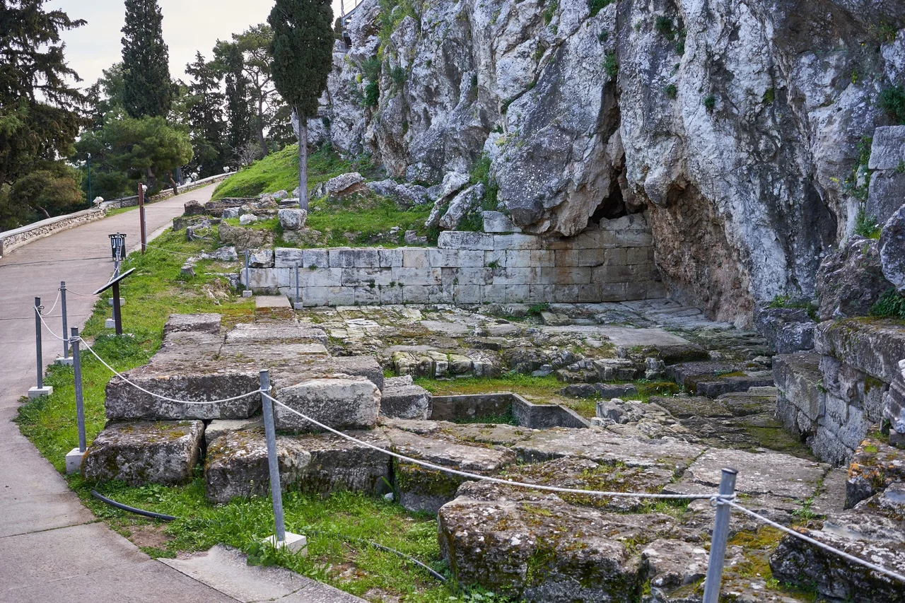 "Klepsydra is the most important spring on the Acropolis rock. Functional since antiquity, it is situated at a nodal point of the North Slope, where the ancient Panathenaic Way meets the Peripatos road, just below the Sacred Caves (of Pan, Zeus and Apollo). The Klepsydra monument is composed of two buildings: the fountain, and the paved court, both built during the second quarter of the 5th cent. B.C. The paved court of external dimensions 24.25 x 10.50m, is situated east of the fountain, and its function is not yet clear: some scholars connect it with the overlying Sacred Caves and suppose that it served as court for the sanctuary of Apollo, while others identify it as a cistern collecting rainwater from the Acropolis...During the 3rd cent. A.D. the court was abandoned and covered with dirt." Information label on the archaeological site.
