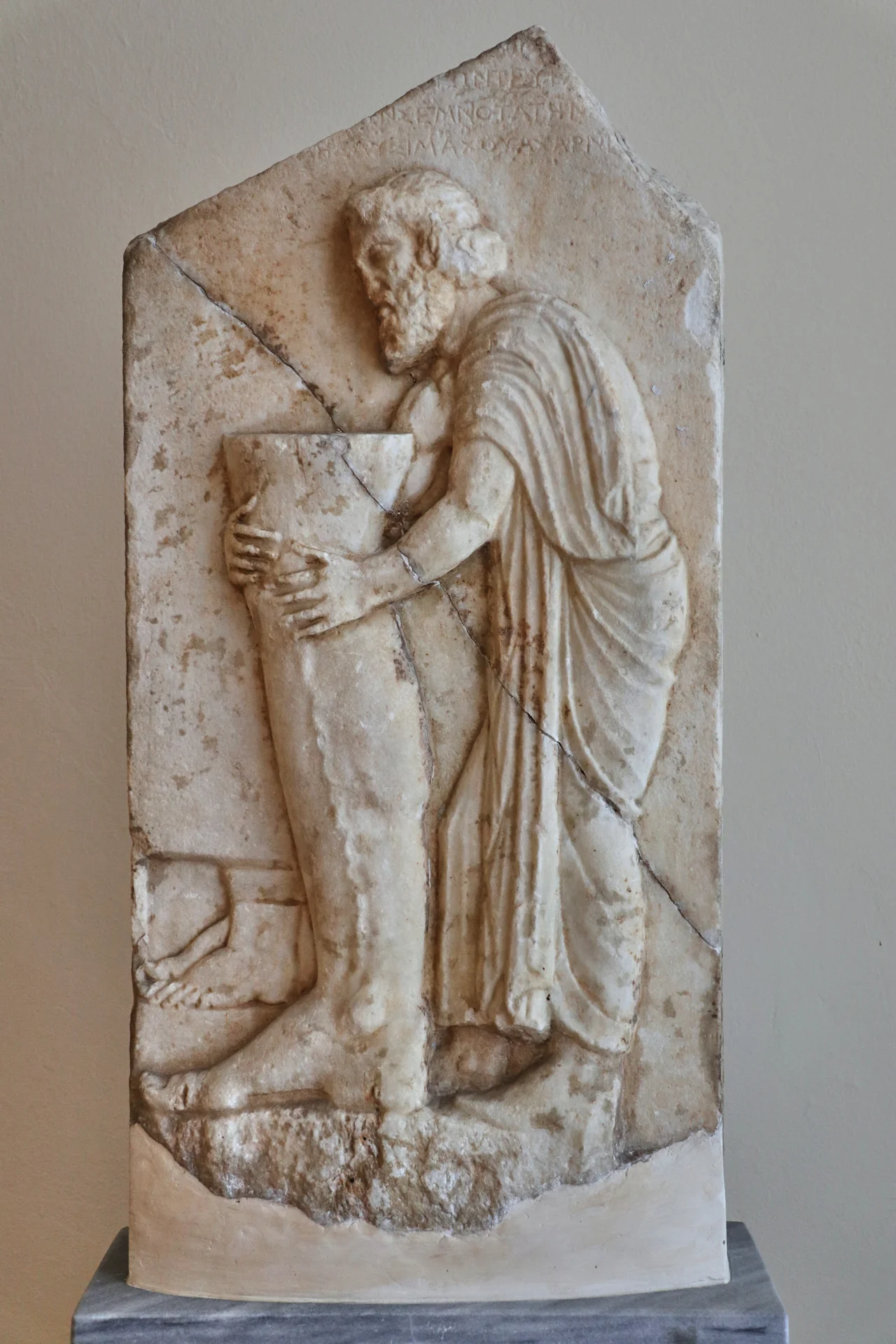 Votive relief. Marble. Found near the Enneakrounos fountain, Athens. From the sanctuary of the hero-physician Amynos. According to the inscription above the representation the relief was dedicated by Lysimachides, son of Lysimachos, from Acharnai. The dedicator holds a model of a human leg, on which a vein stands out indicating his disease. In a recessed panel at the bottom left are represented two feet. End of the 4th century B.C. Accession number: 3526. National Archaeological Museum of Athens. Athens, Greece.
Text: Museum inscription.