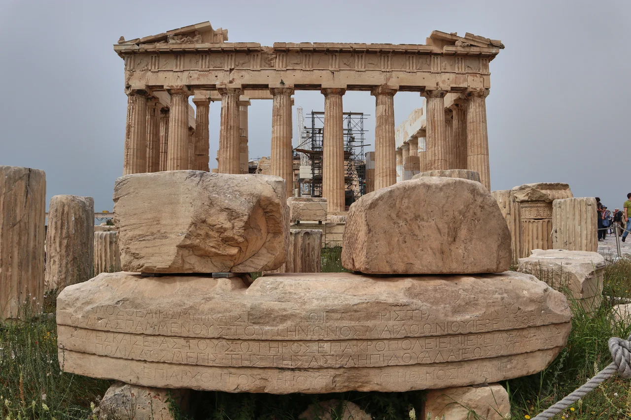 East of the Parthenon lay the foundations of a small building attributed by the first excavators of the Acropolis to the Temple of Rome and the Roman emperor Octavian Augustus. The association of the foundations with the temple stems from the discovery in the area of many marble architectural members, as well as of the architrave bearing the incised dedicatory inscription. (“The deme (dedicated) to the goddess Rome and Sebastos Caesar (Augustus), when general of the hoplites was Pammenes, son of Zenon from Marathon, the priest of the goddess Rome and Sebastos Soter on the Acropolis; when priestess of Athena Polias was Megiste, daughter of Asclepides from the deme of Halai. When Eponymous Archon was Areos, son od Dorion from Paiania.”)
The architectural members indicate that the Temple of Rome and Augustus was of the Ionic order, circular and monopteral – namely that it featured a single circular colonnade made of nine columns (pteron), without a walled room inside (cella). Its diameter measured ca. 8.60 m. and its height reached 7.30m. up to the conical roof. The construction of the temple is associated with the architect who repaired the Erechtheion in the Roman Period, because the architectural details of its members replicate those of the Erechtheion. It is possible that the temple interior housed statues of Rome and Augustus, although no fragments have been identified to date.
The temple of Rome and Augustus is the sole Roman temple on the Acropolis and the only Athenian temple dedicated to the cult of the Emperor. The Athenian deme (people) constructed it in order to propitiate Octavian August and reverse the negative climate that characterized the relations of the two parties , as, during the Roman civil wars, the city of Athens had supported his opponent, Marcus Antonius.
The temple is securely dated after 27 B.C., when Octavian was proclaimed Augustus – most probably between 19 and 17 B.C.
Text credit: Inscription at the archaeological site.