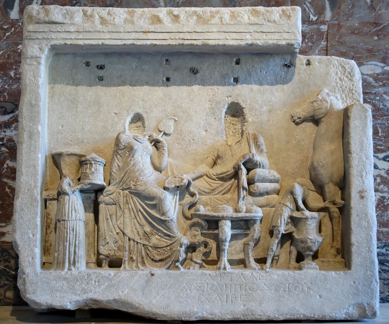 Funerary banquet: from left to right, a servant holding a round object, perhaps a model of the Arsinoeion in Samothrace, a seated woman, a half-reclining man holding a phiale in which a snake is drinking, a boy cupbearer. Naiskos-style funerary stele with high-relief decoration ; epitaph inscribed on the plint: “Attalos, son of Asklepiodoros, greetings!”. Marble, 2nd quarter of the 2nd century BC. From Cyzicus.