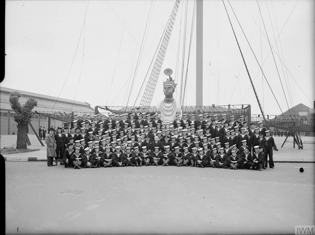 Mr W J Jordan, the New Zealand High Commissioner, with a very large group of New Zealand sailors at the naval training establishment HMS GANGES, Shotley. They are stood in front of the masthead and figurehead of HMS GANGES.