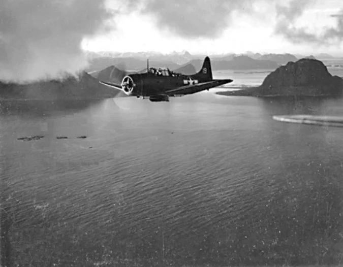 A Douglas SBD Dauntless of U.S. Navy bombing squadron VB-4 during Operation Leader , on 4 October 1943, flying from the aircraft carrier USS Ranger (CV-4). The objective of the force was the Norwegian port of Bodø. The task force reached launch position off Vestfjord before dawn 4 October completely undetected. At 0618, Ranger launched 20 Dauntless dive bombers and an escort of eight Grumman F4F-4 Wildcat fighters of VF-4. One division of dive bombers attacked the 8,000-ton freighter LaPlata, while the rest continued north to attack a small German convoy. They severely damaged a 10,000-ton tanker and a smaller troop transport. They also sank two of four small German merchantmen in the Bodø roadstead.
The SBD "4-B-19" was piloted by Lt(jg) Clyde A. Tucker with ARM2c Stephen D. Bakran as gunner and passes Kunna Head South of Bodø. It was one of two planes later lost in the attack.