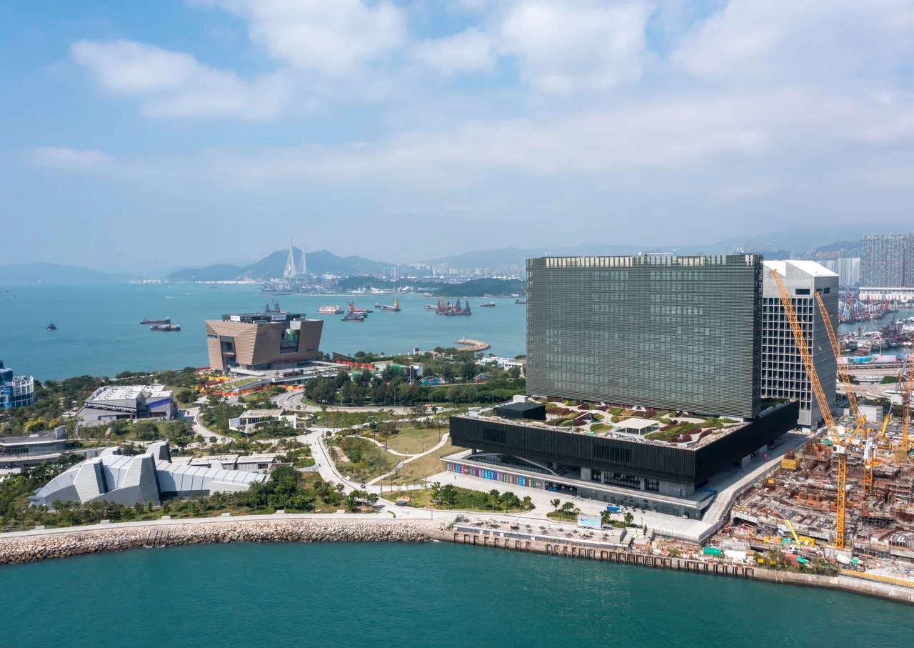 An aerial view of M+ in West Kowloon, Hong Kong. A construction site sits to the right of the museum. To the left is a green, leafy park. The Palace Museum is visible in the background. Victoria Harbour lies in the foreground of the scene.